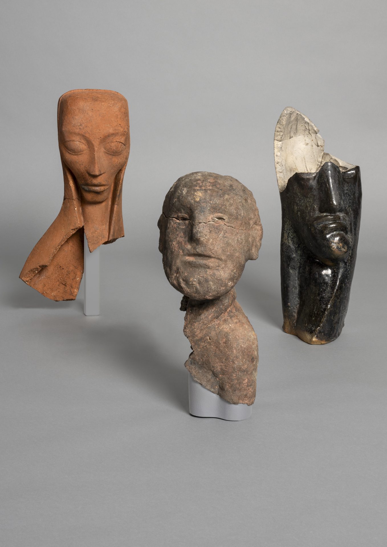 Three artistic sculptures of heads in various shapes and materials against a gray background.